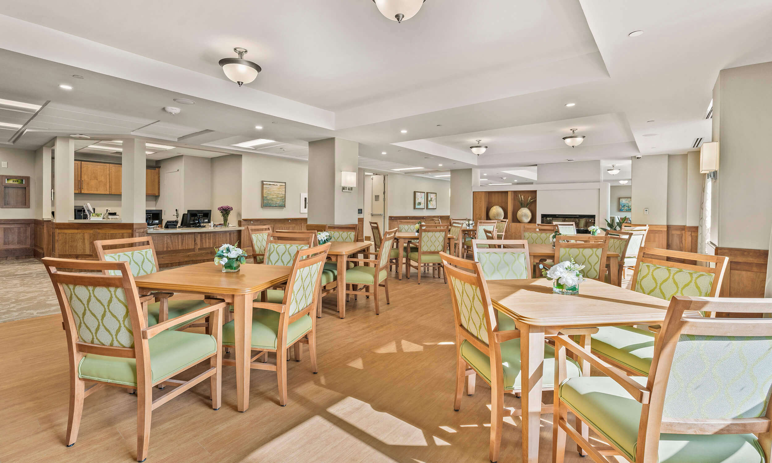 Dining room of retirement home with eight wooden set tables and chairs with green cushioning