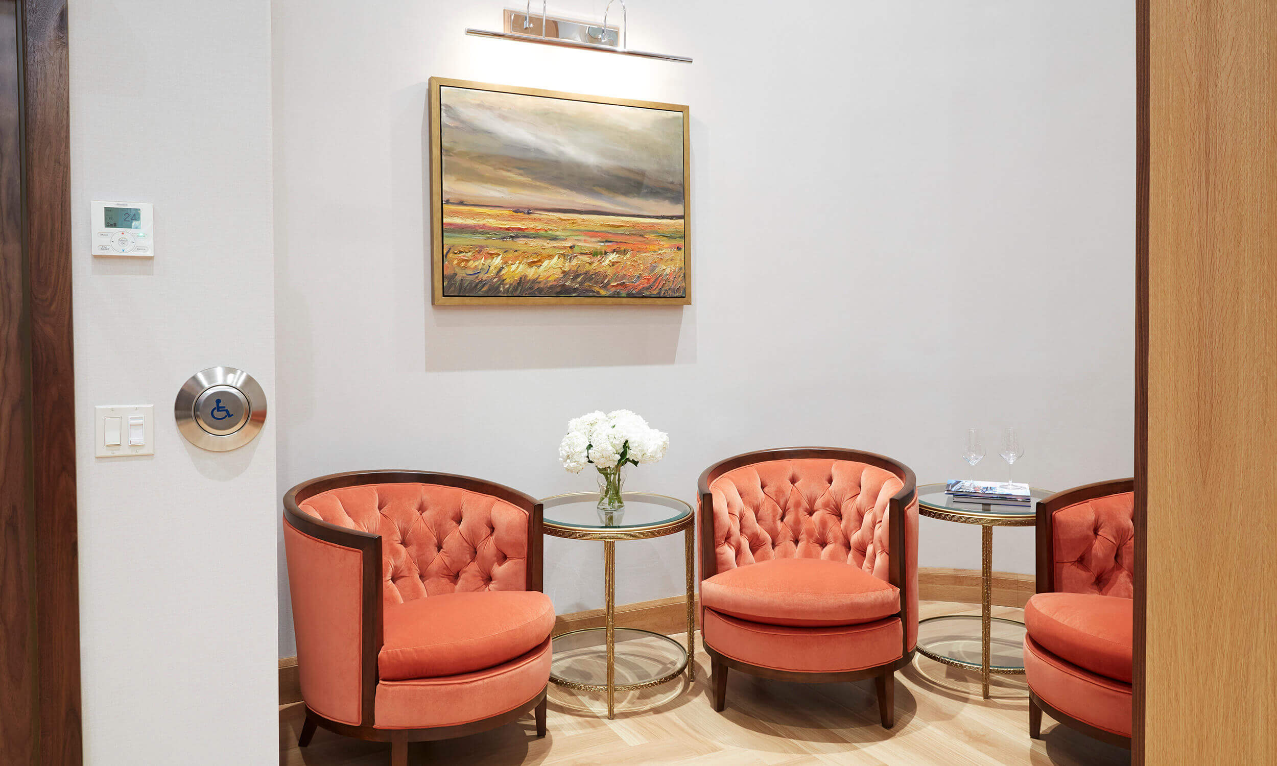Waiting room in retirement home with three orange plush chairs with small tables between and landscape painting on wall