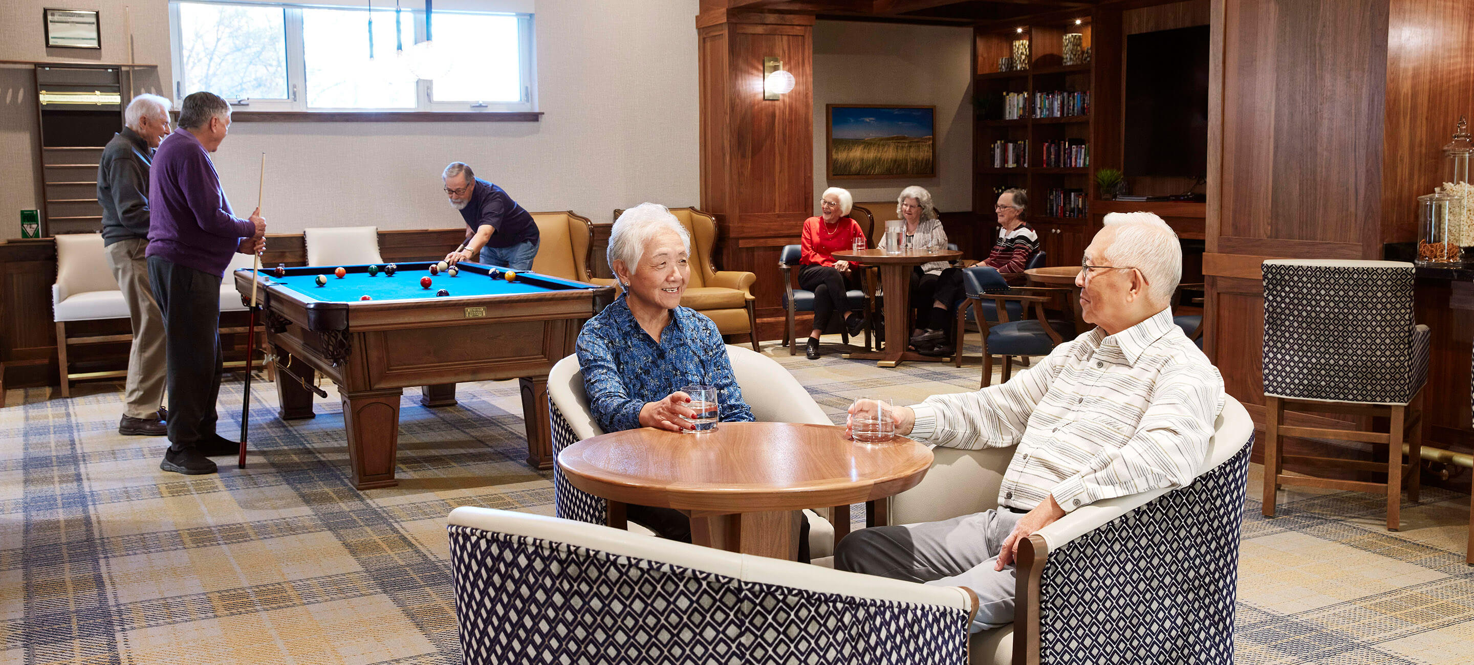 Entertainment room in retirement home with guests sitting at tables holding drinks while others play pool