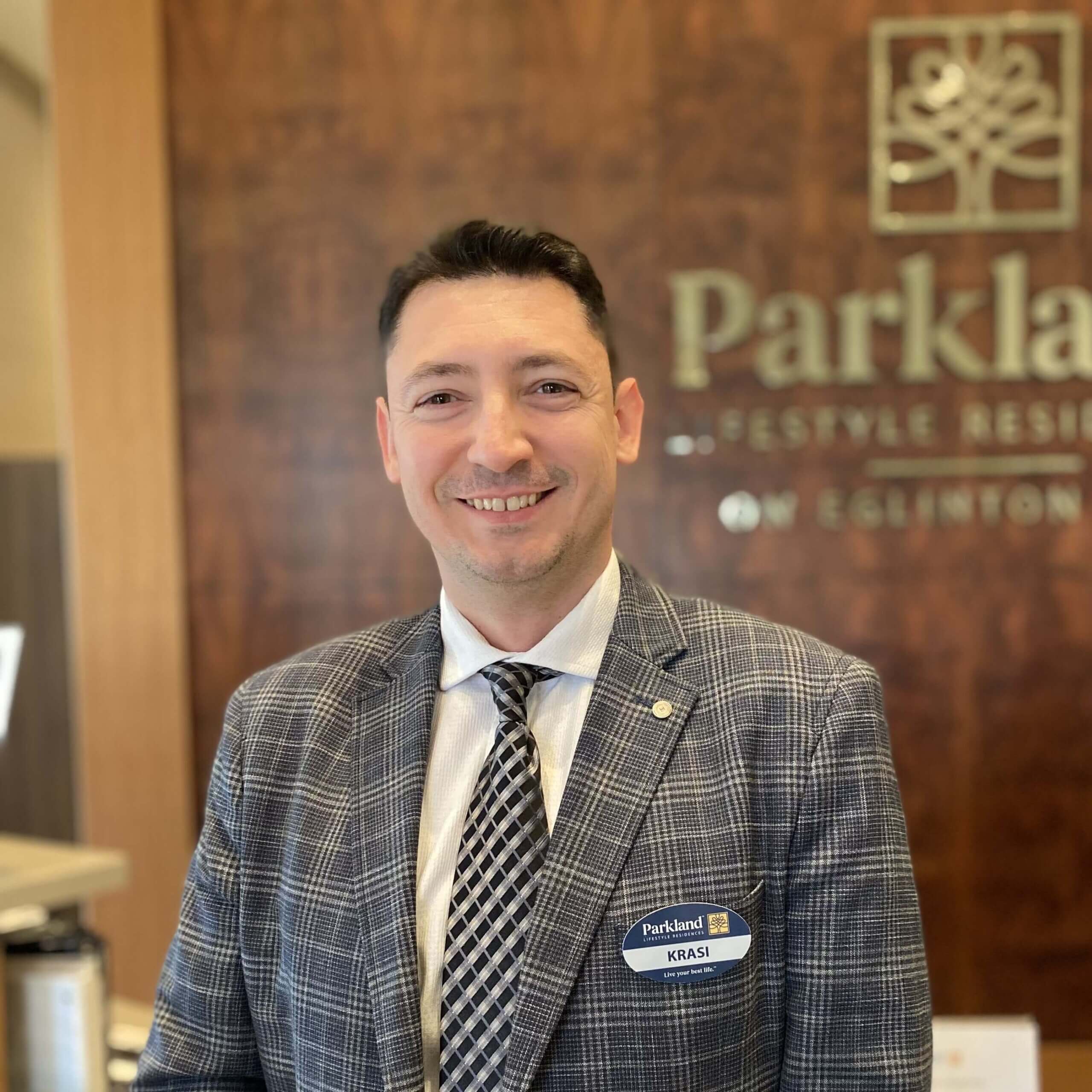 a person smiling in a professional setting. They are wearing a gray checkered blazer, a light-colored dress shirt, and a patterned tie. A name badge is visible on their blazer, which has a company logo. The background includes a wooden panel with a company name, suggesting a corporate or hospitality environment.