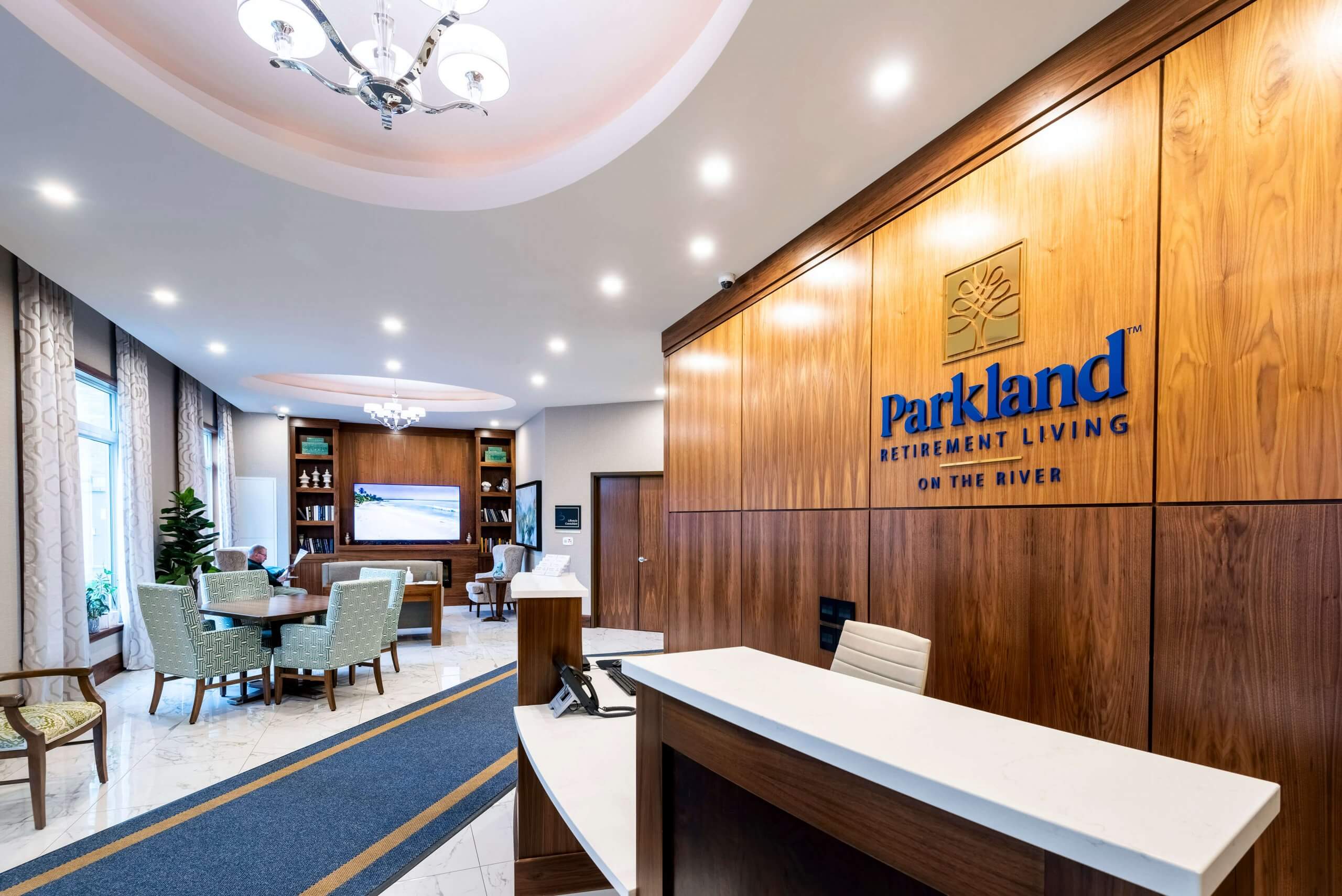 Lobby of Parkland Residence with wooden front desk in front of Parkland sign on wall and surrounding furniture with table and television