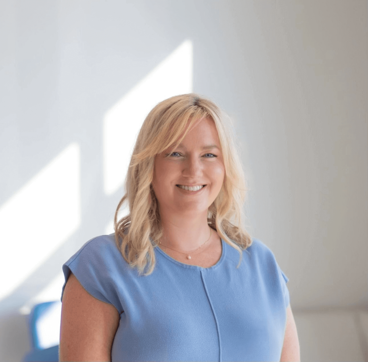 A person with shoulder-length blonde hair is smiling and wearing a light blue top in a well-lit room with a plain background.