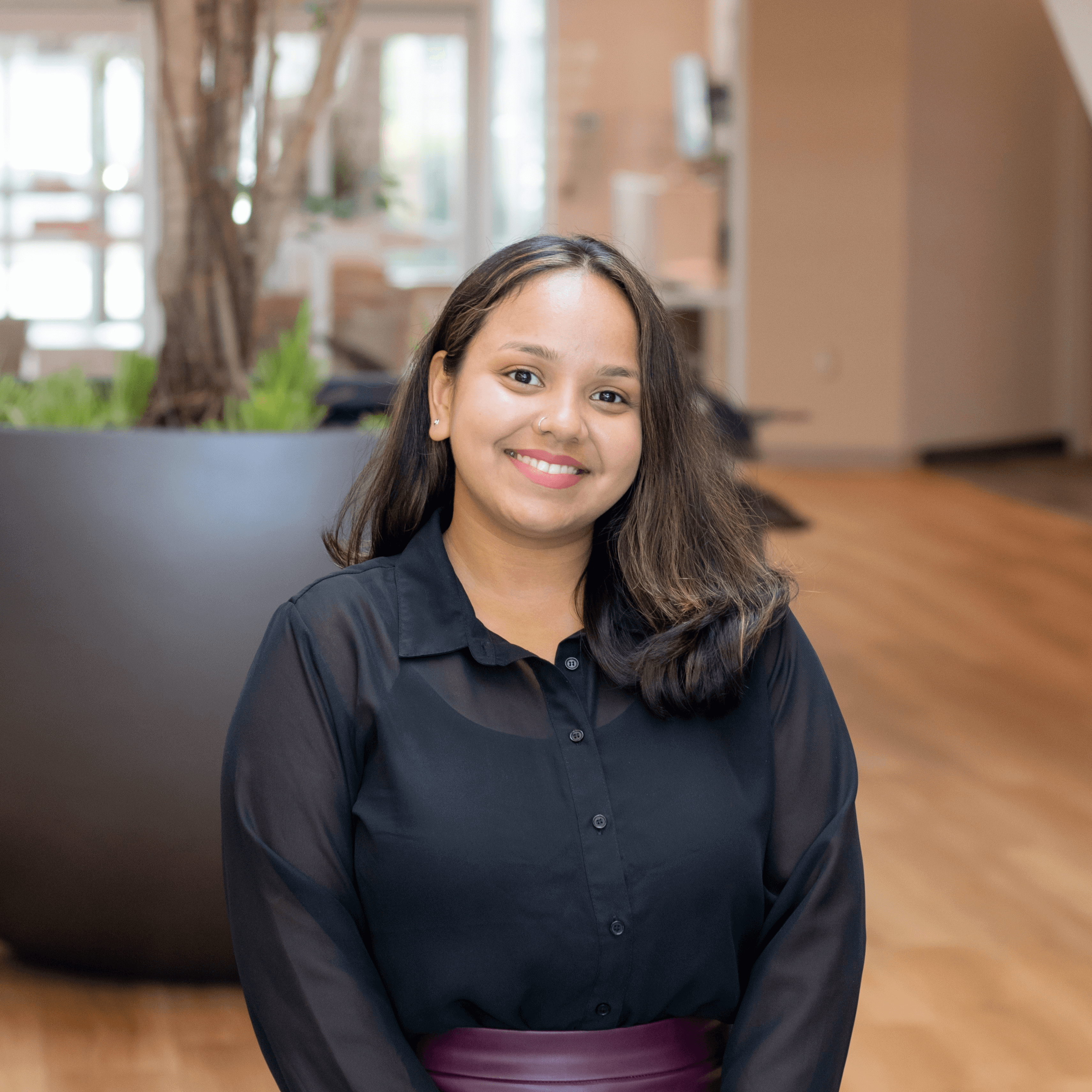 a person standing indoors with a friendly smile. They have medium-length, dark hair with light highlights, and are wearing a black blouse. The background includes indoor plants and wooden flooring, suggesting a modern, comfortable environment.